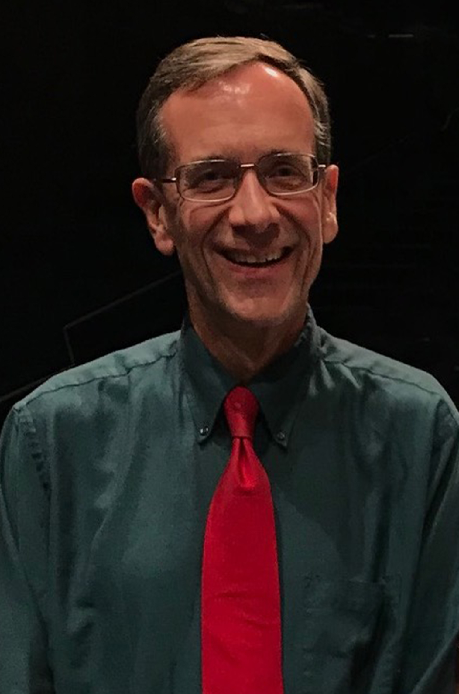 Matt Stock has short brown hair and glasses and is smiling. He is wearing a dark green, collared shirt and a bright red tie against a dark background.